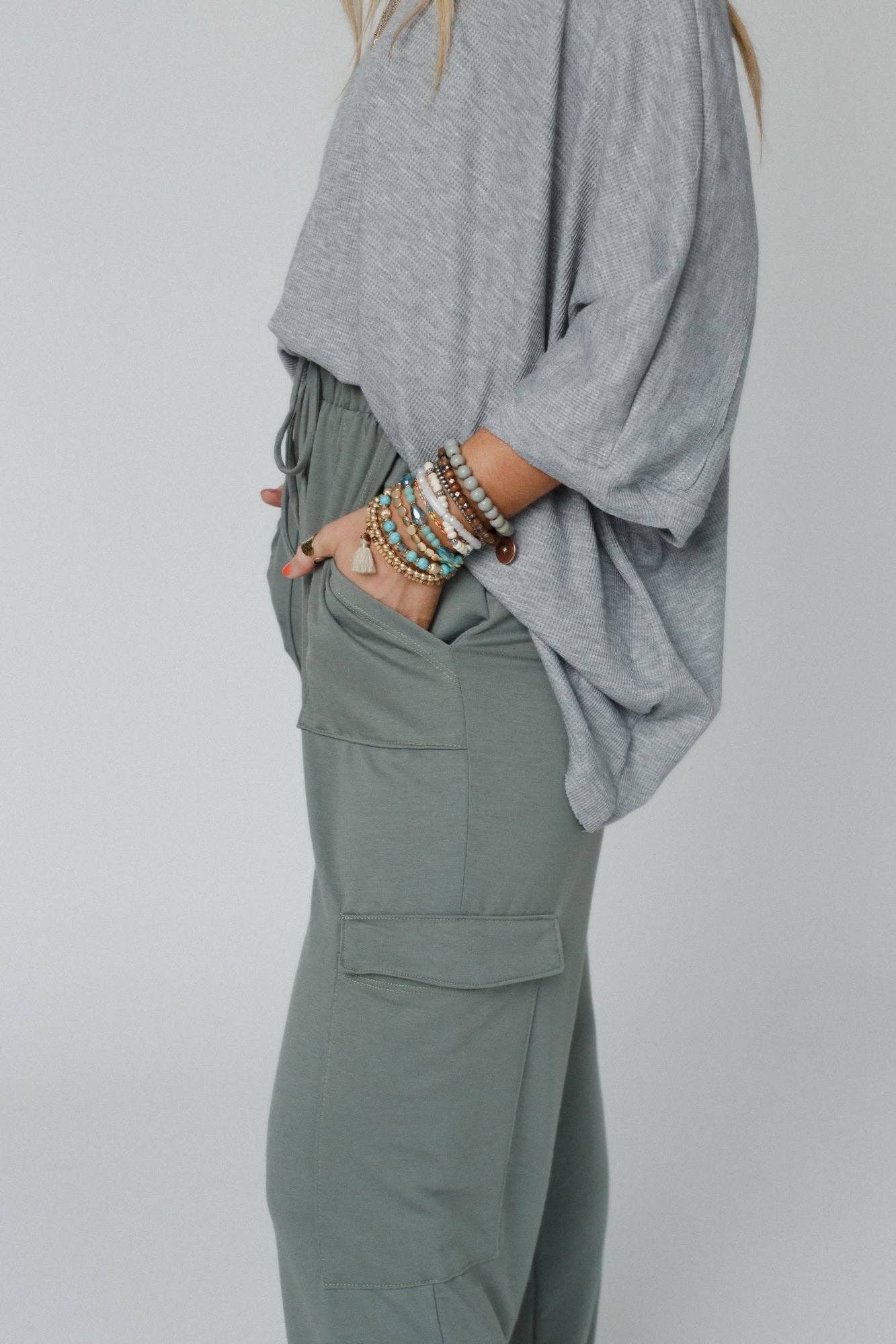 Side view of the Easy Mornings Cinched Pants by Three Bird Nest in light olive, styled with a loose gray top and bracelets.