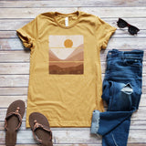 A yellow graphic tee with a sunset design, displayed flat with a pair of blue jeans, brown sandals, and accessories.