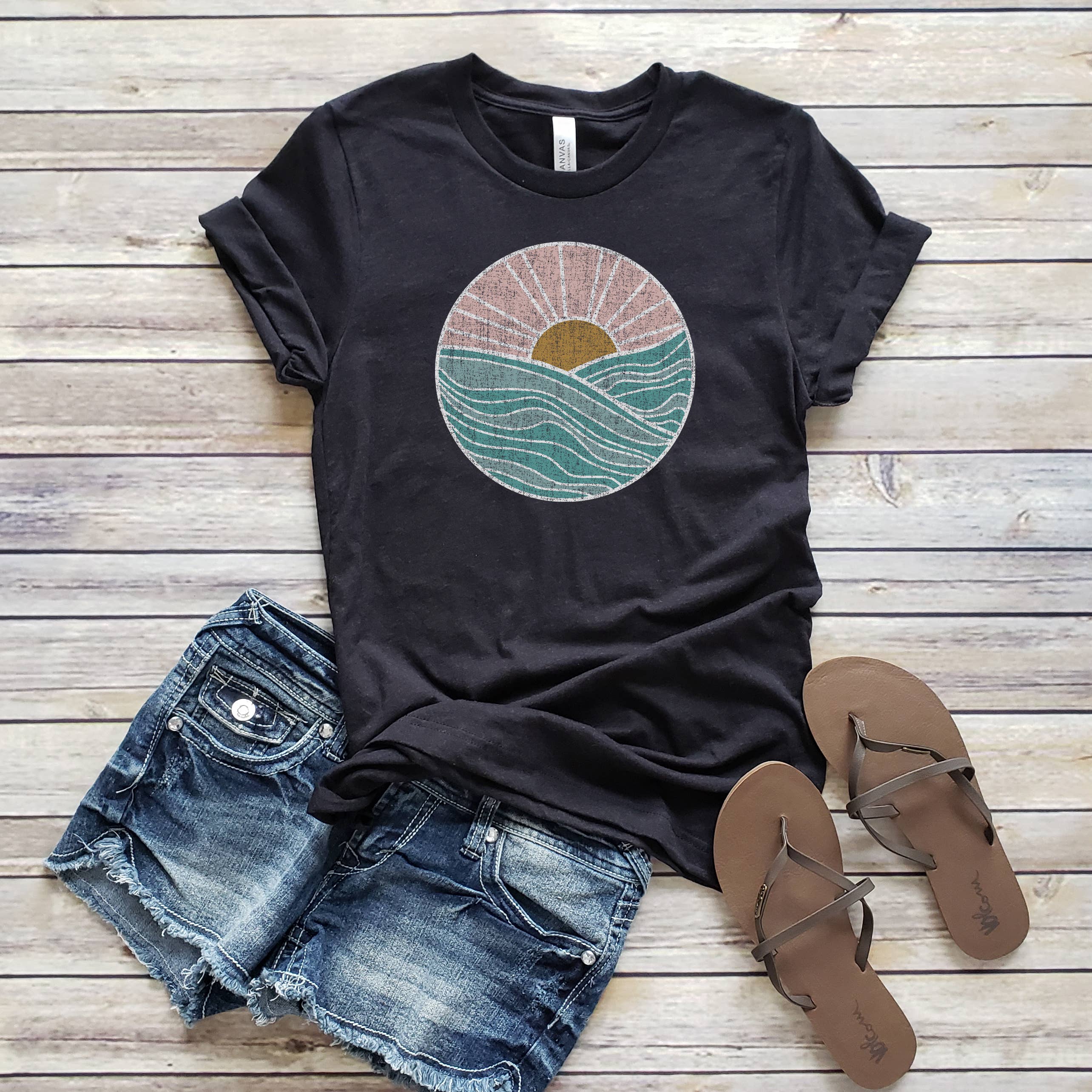 A black t-shirt with a circle sunset graphic design, accompanied by denim shorts and a pair of sandals on a wooden surface.