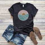 A black t-shirt with a circle sunset graphic design, accompanied by denim shorts and a pair of sandals on a wooden surface.