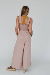 Back view of the Three Bird Nest Smocked Ruffle Jumpsuit in dusty rose, showing ruffle details and flowing fabric, styled with loose hair and a casual summer vibe.