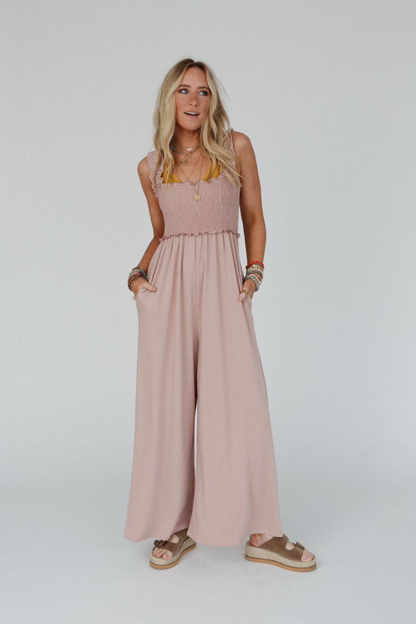 Model wearing the Three Bird Nest Nothing Compares Smocked Ruffle Jumpsuit in dusty rose, shown from the front with smocked top and flowing wide-leg pants, styled casually with a straw hat and neutral-toned sandals.