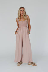 Model wearing the Three Bird Nest Nothing Compares Smocked Ruffle Jumpsuit in dusty rose, shown from the front with smocked top and flowing wide-leg pants, styled casually with a straw hat and neutral-toned sandals.