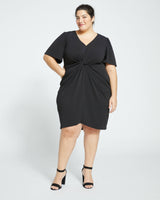 A woman wearing a black knee-length dress with flutter sleeves and a twist detail under the bust. She is paired with black ankle strap sandals.